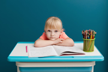 Portrait of little blonde girl is sitting at the table and paints. Schoolkid is doing her homework in classroom at school or at home. Tired and sad pupil