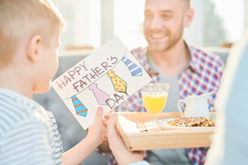 Warm toned shot of little boy giving his dad handmade Fathers day card and surprise breakfast at home, copy space