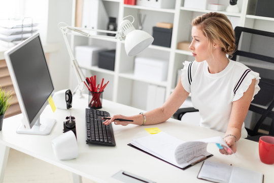 A young blonde girl is sitting at a computer desk in the office, holding a pencil in her hand and working with documents.