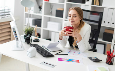 A young girl is sitting at the computer desk in the office and holding a cup. Before the girl on the table are diagrams.