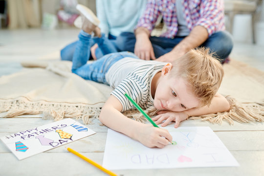 Warm toned portrait of cute little boy lying on floor in living room drawing picture for Fathers day for dad and smiling, copy space