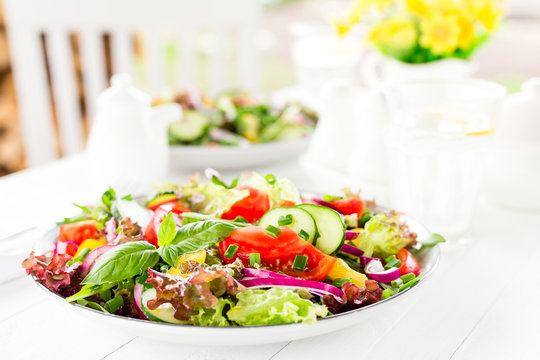 Fresh vegetarian vegetable salad with tomato, onion, cucumber, pepper, basil and lettuce. Salad on plate on white table