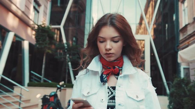 Beautiful, curly young woman in a stylish outfit with trendy scarf on her neck walks down the city street while using her smartphone. Modern technologies, surfing the internet, online chatting.