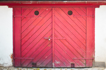 The gates are made of wood and painted red and mounted on a white wall. The gate doors are closed with a metal padlock. Red background of wooden planks.