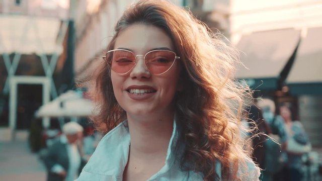 Pretty young curly woman looking straight to camera in cheerful mood with a happy smile, wearing stylish hipster sunglasses, spring summer fashion trend, denim jacket. Having fun, date, joyful mood.