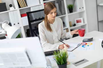 A young girl is sitting at a table in the office, holding a red cup in her hand and writing in a notebook with a pencil.