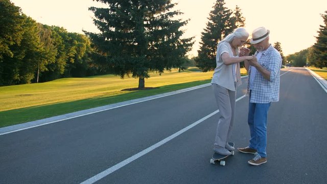Happy retired couple having fun skateboarding in summer park. Beautiful aged woman with gray hair practiving and learning to skateboard while husband teaching her during sunset. Steadicam shot