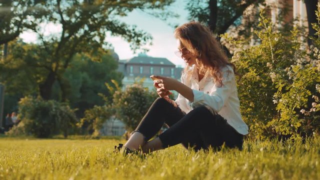 Low angle shot of pretty curly young woman in stylish white jacket and sunglasses sitting on the grass in the city park, under the blossoming bushes, and using her smartphone