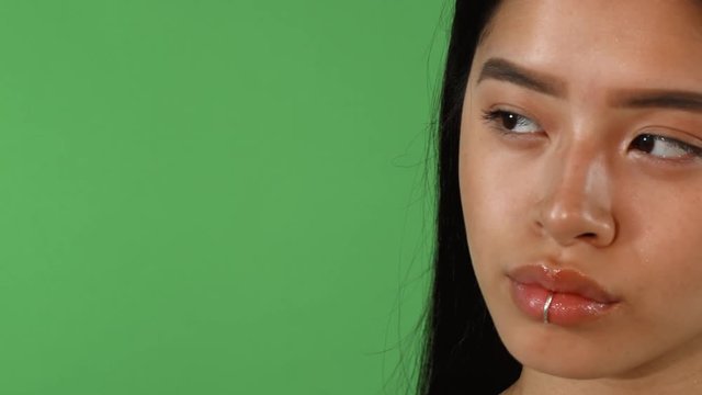 Sliding cropped shot of a gorgeous young Asian woman with flawless skin and lip ring piercing posing confidently on green chromakey background, looking away. Copy space on the side.