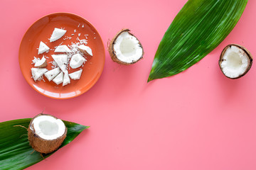 Fototapeta na wymiar Pieces of coconut flesh on plate on pink background with coconut cut in half and palm leaves top view space for text