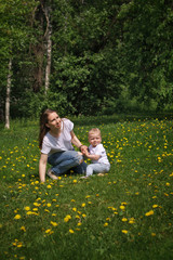 Fototapeta premium Family. Mom and son in meadow. Girl and little boy are sitting on green lawn. Yellow dandelions growing in meadow.