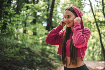 Fototapeta premium Beautiful sports woman put on headphones on training outdoors. Young athletic woman in pink hoodie put on headphones before running in the forest. Portrait of sports smiling girl listening the music.