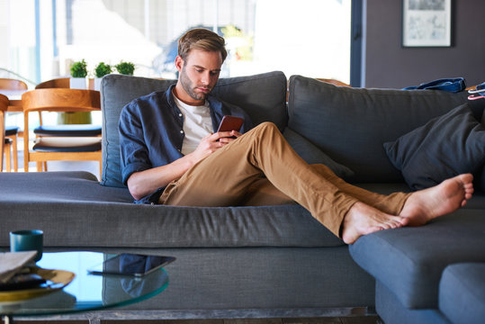 Wide shot of an attractive caucasian man sitting on a generous sofa in a modern home, while texting on his latest mobile phone with his feet up on the couch and relaxing.