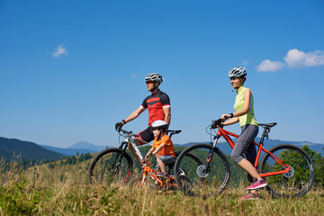 Young family tourist bikers, mom, dad and child resting with bicycles, looking into distance, standing on grassy hill on summer sunny day. Active lifestyle, traveling and happy relations concept.