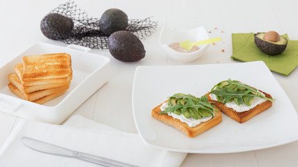 Roasted toasts with cream cheese and slices of avocado, leafs of rucola and pink Himalayan salt. Front view, low angle, focus on the centre of both breads. White wood background.