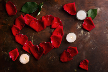 Red rose petals in heart shape with white candles on dark brown and golden background. Love, romance, anniversary, valentine's day concept. Text space