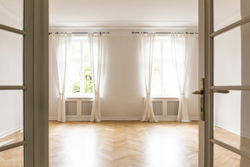 Empty bright living room interior with drapes at windows and wooden floor. Real photo