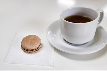A cup of tea on a white saucer with brown macaroon on a white table.