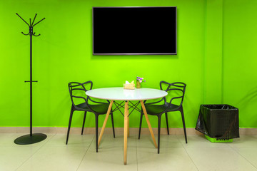 Interior in bright green colors with a table, two chairs, a hanger, a trash can and a TV.
