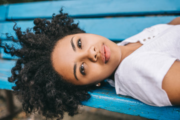 Attractive african american woman having rest outdoor on bench in the park, looking at the camera. Beautiful young dark-skinned female wears white t-shirt enjoy healthy life. People, lifestyle concept