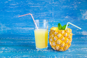 A glass of pineapple juice with a straw and a cut pineapple with mint leaves on a blue rustic background.
