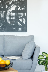 Real photo of a grey sofa standing next to a plant and behind a table with fruit in living room interior with black and white painting on a wall