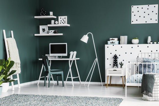 Dark green teenager room interior with a desk, chair, computer, lamp, shelves, decorations and bed set on a creative wall