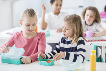 Snack time in a kindergarten class. Children opening their mint color lunch boxes with healthy vegetables. A bottle of orange juice on the desk.