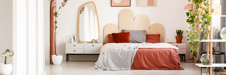 Bright bedroom interior with king-size bed with grey and red bedding, white cupboard with decor,...