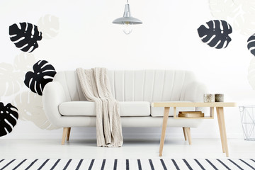 Wooden table in front of settee with blanket in white flat interior with monstera leaves. Real photo
