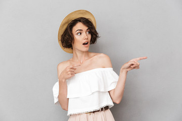 Excited european woman 20s wearing straw hat looking aside and pointing finger at copyspace with open mouth, isolated over gray background