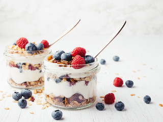 Two jars with tasty parfaits made of granola, berries and yogurt on white wooden table. Shot at...
