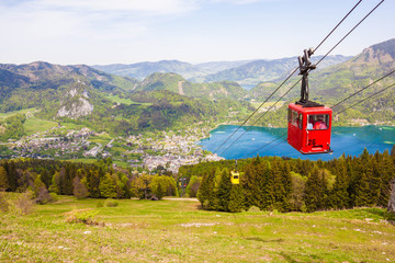 Yellow and red gondolas of Zwoelferhorn Seilbahn (cable way) travel up and down alpine peak with a view of village St.Gilgen, Wolfgangsee lake, surrounding hills and mountains. Salzburg land, Austria - obrazy, fototapety, plakaty