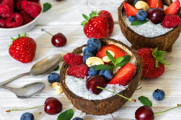 Vegan chia seeds pudding with fresh berries, nuts and mint in coconut bowls with spoons over white wooden table