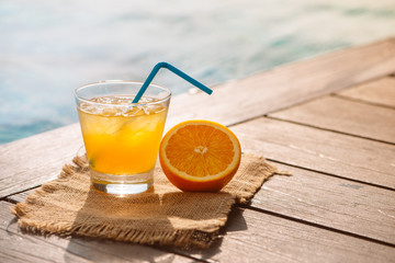 Close up of screwdriver cocktail alcohol drink with orange juice, slices and ice standing near the pool. Refreshing iced lemonade beverage in glass by the poolside. Sun glares. Background, copy space.
