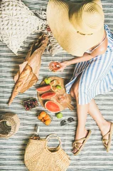 Foto op Plexiglas Picknick Zomer picknick instelling. Vrouw in linnen gestreepte jurk en stro zonnehoed met glas rose wijn in de hand, vers fruit en stokbrood op deken, bovenaanzicht. Buitenbijeenkomst of lunch