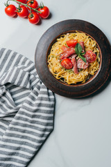 elevated view of pasta with mint leaves, jamon and cherry tomatoes covered by parmesan on plate at marble table with kitchen towel