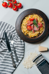 top view of pasta with mint leaves, jamon and cherry tomatoes covered by parmesan on plate at marble table with kitchen towel, knife, fork, cheese and grater