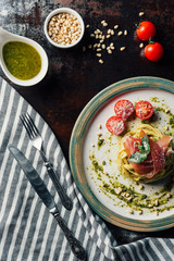 top view of pasta with mint leaves, jamon and cherry tomatoes covered by parmesan on plate at table with kitchen towel, knife, fork, pine nuts and pesto in bowl