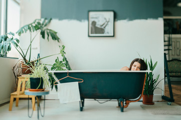 Tilt shift portrait of young beautiful girl in pink dress lying in empty vintage cast-iron bath inside decorative in french style room with green plants. Odd unusual strange woman home relaxation.
