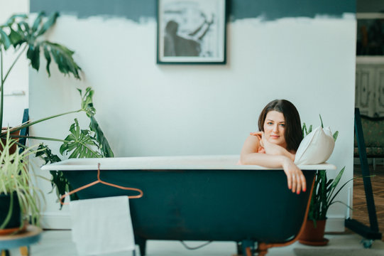 Tilt Shift Portrait Of Young Beautiful Girl In Pink Dress Lying In Empty Vintage Cast-iron Bath Inside Decorative In French Style Room With Green Plants. Odd Unusual Strange Woman Home Relaxation.