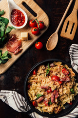 elevated view of pasta with jamon, pine nuts, sauce, cherry tomatoes, mint leaves covered by grated parmesan in pan surrounded by kitchen towel and cutting board with ingredients on table