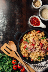 elevated view of pasta with jamon, pine nuts, sauce, cherry tomatoes, mint leaves covered by grated parmesan in pan surrounded by ingredients and kitchen towel on table