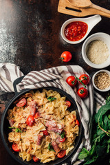 top view of pasta with jamon, pine nuts, sauce, cherry tomatoes, mint leaves covered by grated parmesan in pan surrounded by ingredients and kitchen towel on table