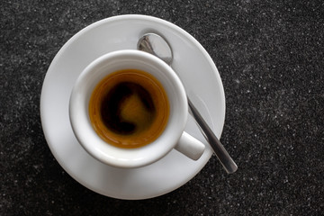 Small italian espresso in white ceramic cup with spoon isolated on dark granite desk from above.