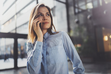 Charming young business woman talking with partner via mobile phone while standing at modern business center.