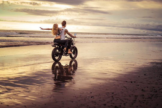 girlfriend with open arms sitting on motorcycle after boyfriend on ocean beach