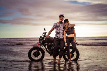 Obraz premium stylish boyfriend hugging girlfriend near motorcycle on ocean beach and they looking at camera