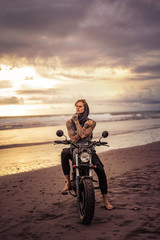 Fototapeta na wymiar pensive handsome tattooed biker sitting on motorcycle on ocean beach