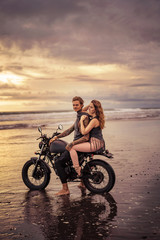 Fototapeta premium side view of boyfriend and girlfriend sitting on motorcycle at beach during sunrise